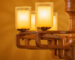 Close-up of a modern wooden chandelier with geometric arms and cylindrical frosted glass shades, radiating warm ambient light against a golden background